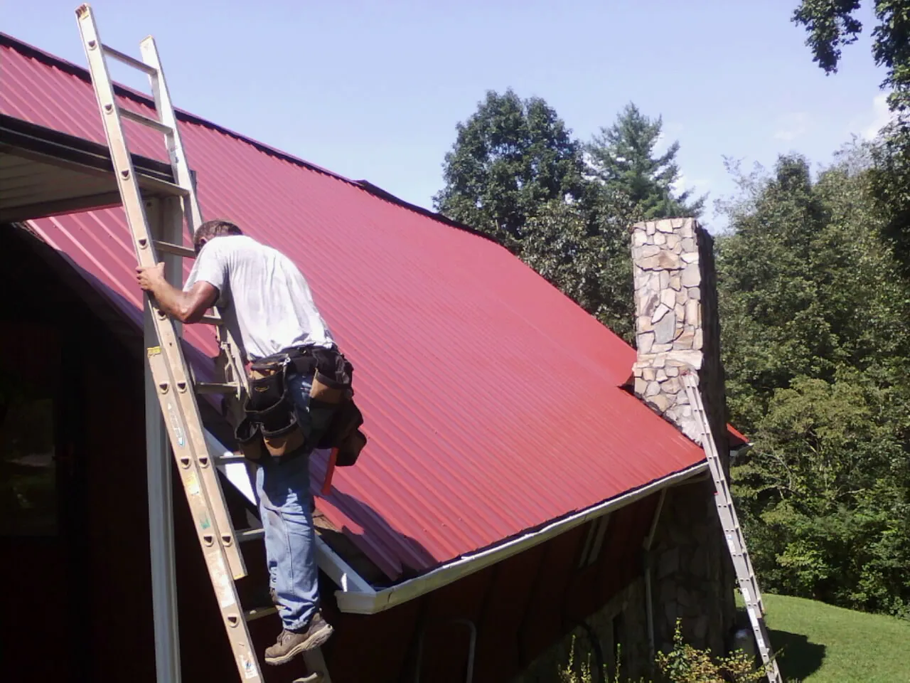 Roofer on a ladder beside a red metal roof and stone chimney — Soffit & Fascia Repair by Roofing Brothers in Eatontown
